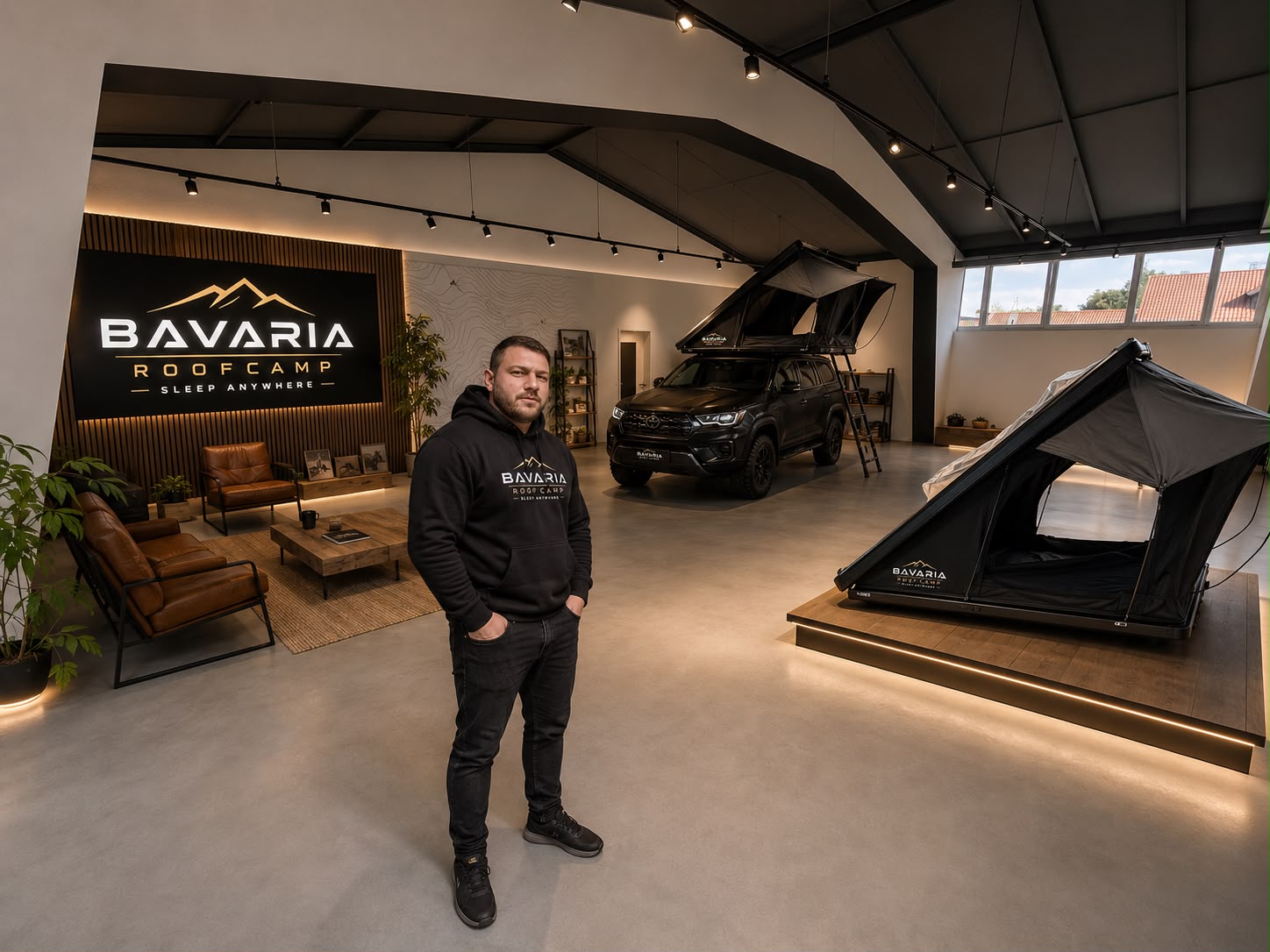Der Inhaber steht in der Ausstellungshalle von Bavaria Roofcamp, dahinter ein Toyota mit aufgebautem Dachzelt und ein zweites Zelt auf einem Holzpodest.