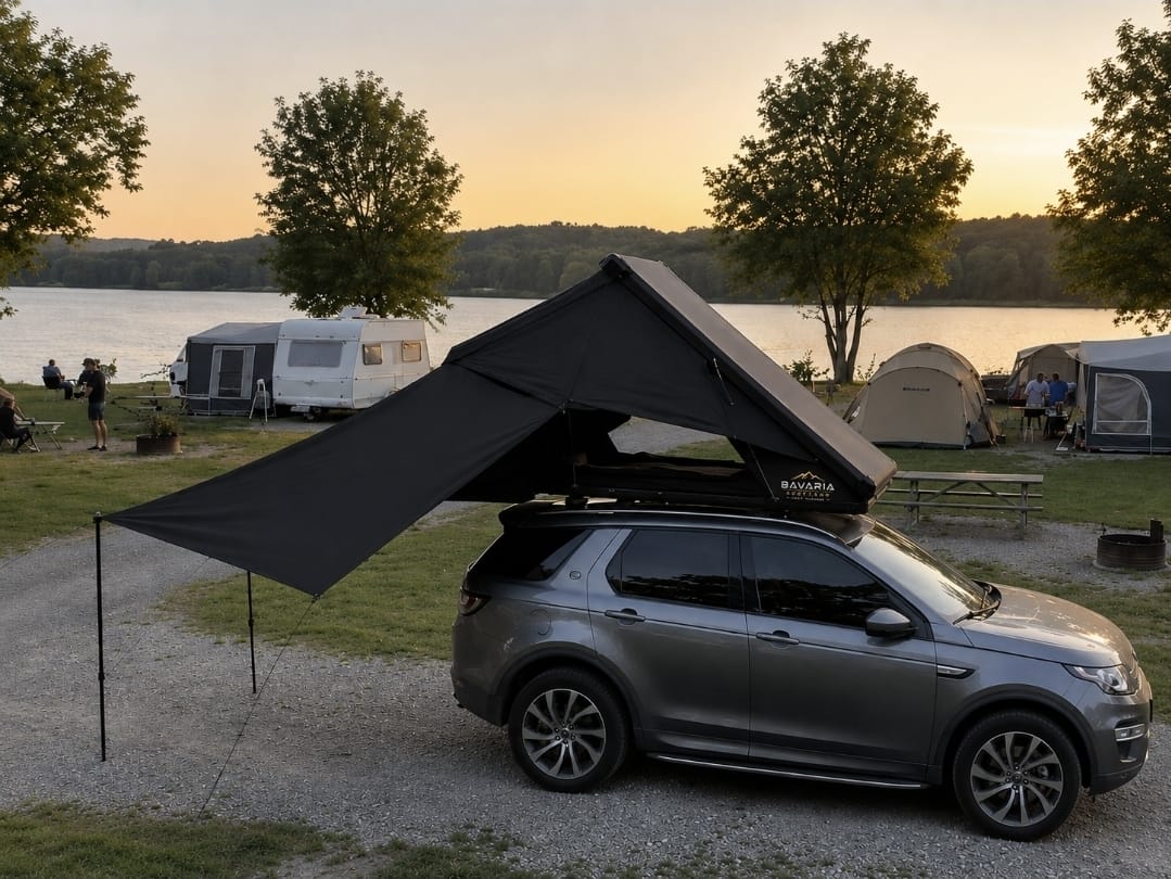 Ein Land Rover Discovery mit Dachzelt und weit aufgespanntem Sonnensegel steht abends auf einem Stellplatz am See.