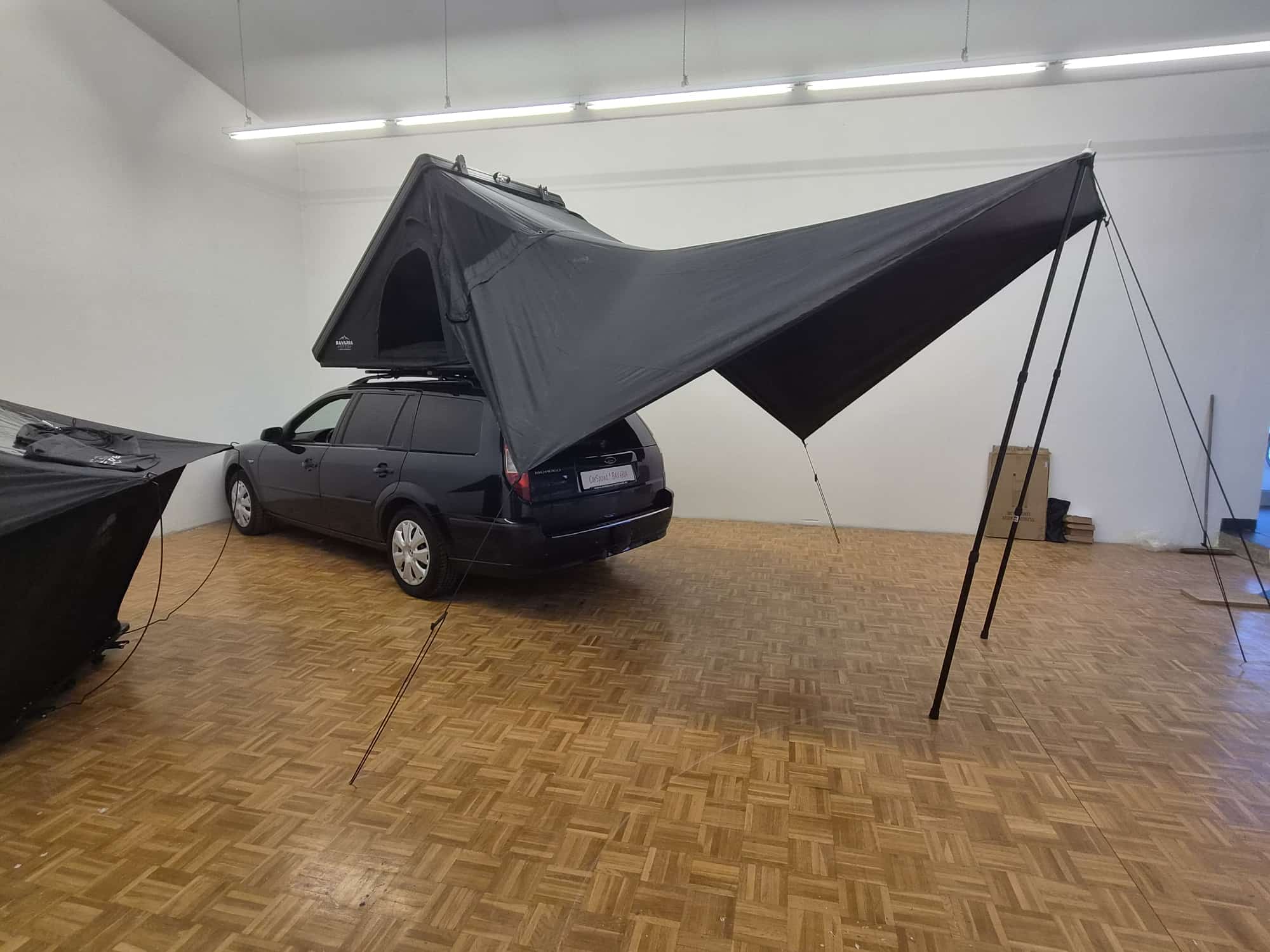 Der Kombi mit aufgebautem Dachzelt, daneben ein zweites Zelt am Boden; das Sonnensegel ist weit über den Platz neben dem Fahrzeug gespannt.