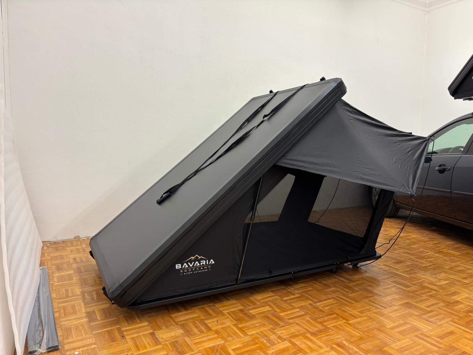 Ein Dachzelt von Bavaria Roofcamp steht aufgeklappt am Boden der Ausstellungshalle, die graue Hartschale weit geöffnet, das seitliche Segel eingehängt.